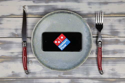Close-up-of-mobile-phone-screen-with-logo-lettering-of-food-delivery-service-dominos-pizza-on-wood-table-with-dish